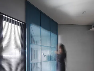 A modern minimalist bedroom featuring a large window with closed blinds, a blue perforated metal panel wall, and a blurred hu