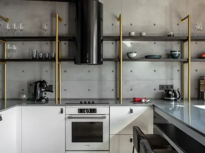 A modern minimalist kitchen with concrete walls, featuring a black and gold shelving unit, white cabinetry, and a sleek oven.