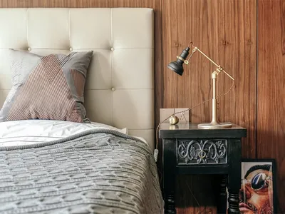 A minimalist bedroom featuring a white leather headboard, a small brass and metal adjustable lamp, and a wooden wall with a f