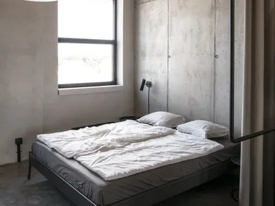 A minimalist bedroom with a concrete floor, white bedding, and industrial-style wood beams on the ceiling.