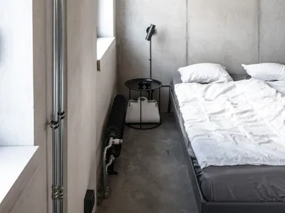 A minimalist bedroom with concrete walls and floor, featuring a white bed with white pillows, a black side table with a black