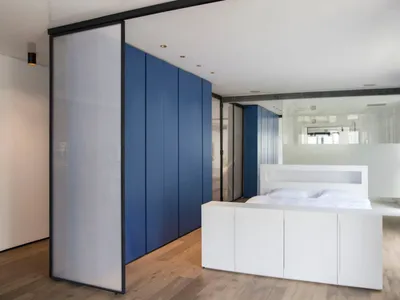 Modern bedroom with white furniture, blue storage cabinets, and glass partition, featuring white floor and wood flooring.