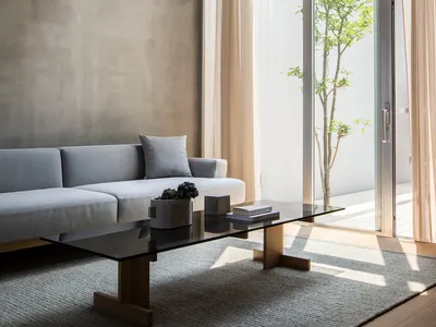 A modern, minimalist living room featuring a concrete wall, glass coffee table, and large floor-to-ceiling windows.