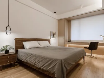 A minimalist bedroom with a wooden bed and a black chair, featuring a large window with white blinds and wood flooring.