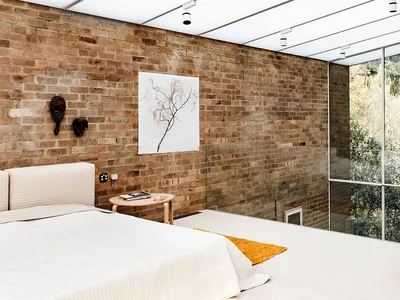 A minimalist bedroom with walls made of brick and concrete, featuring a large window and an orange pouf.