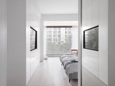 Modern minimalist bedroom with white cabinets and a bed, featuring large windows with a view of urban buildings and greenery.