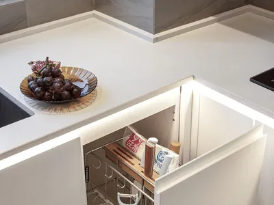A modern, minimalist kitchen with white cabinetry, featuring an organized drawer and a countertop with grapes and a bowl.