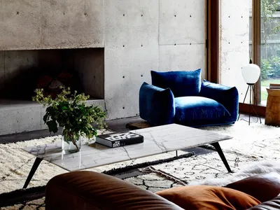 A modern industrial living room with concrete walls, a blue velvet armchair, and a marble coffee table.
