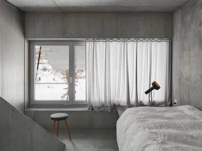 A minimalist bedroom with concrete walls, a white bed, and a white curtain at the window.