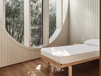A minimalist bedroom featuring a wooden platform bed in front of a large curved window overlooking trees.
