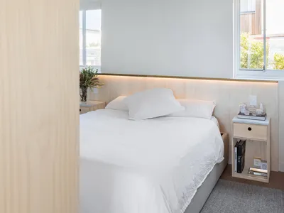 A modern minimalist bedroom featuring a bed with white sheets and a wooden headboard against a white wall, supplemented by am