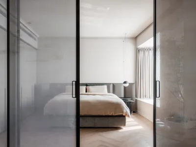 A minimalist bedroom with a modern design, featuring a bed with a cream-colored duvet and a large window letting in natural l