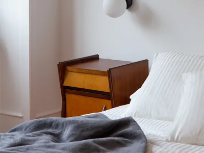 A minimalist bedroom featuring a white wall with a sleek, modern light fixture, a wooden bedside table, and striped bedding.