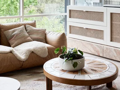 A modern living room with a large window, a tan leather sofa, a round wooden coffee table with a marble top, and a white cabi