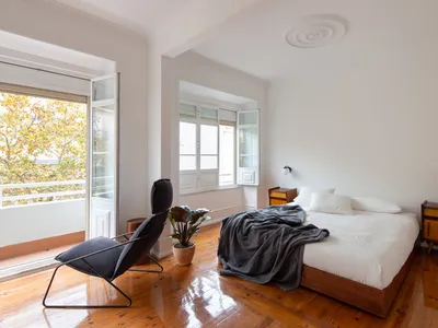 Minimalist bedroom with wooden floors, a bed covered with a gray blanket, and a modern black chair.