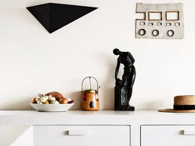 A minimalist kitchen with white cabinets, concrete countertops, a bowl of colorful vegetables, and a black sculpture on the c