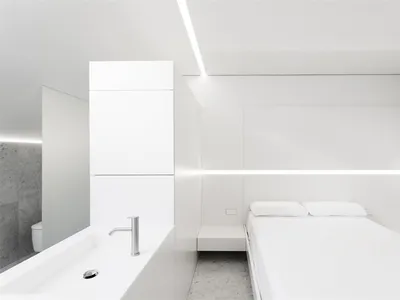 A modern minimalist bedroom with white furniture, a bed with white bedding, and a white sink with a mirror.