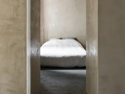 A minimalist bedroom with concrete walls and floor, featuring a white bed and a hanging light bulb.