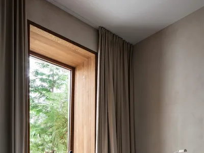 A minimalist bedroom with neutral-colored walls, curtains, and bedding. A small desk with a lamp and chair is placed next to