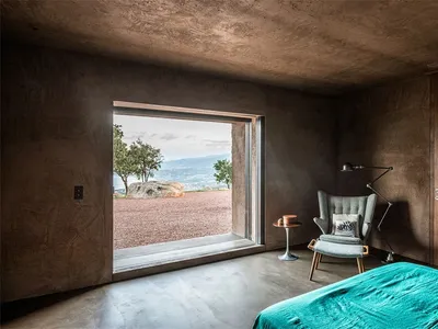 A modern bedroom with a turquoise bedspread, a concrete floor, and a large window overlooking a scenic landscape.