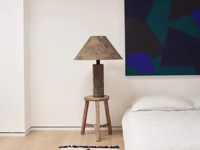 A minimalist bedroom featuring a wooden stool lamp and a white bed.