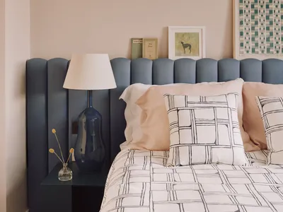 A minimalist bedroom featuring a bed with patterned bedding, a blue headboard, and a side table with a lamp and decorative va