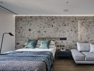 A modern bedroom featuring a concrete wall, a cozy bed with blue and grey bedding, and a comfortable grey couch.