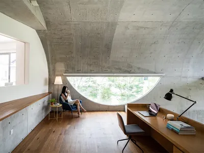 A modern home office features a concrete console with a built-in desk, a woman sitting on a blue armchair by a round window,