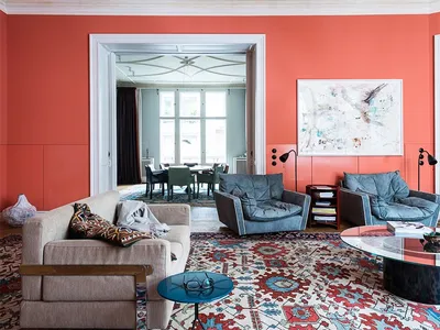 A modern living room with coral-colored walls and a patterned rug, featuring two blue armchairs and a beige sofa.