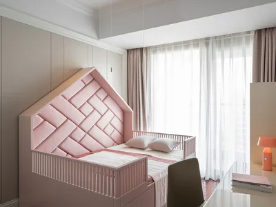 A minimalist bedroom featuring a pink bed with a woven headboard, a grey desk, and a window with sheer curtains.