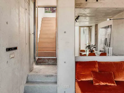 An industrial-style living room with exposed concrete walls and floors, featuring an orange velvet sofa and minimalist decor.