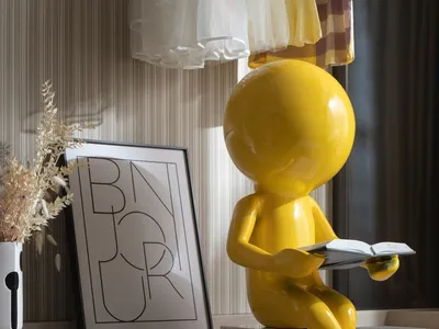 A modern children's bedroom featuring a yellow decorative figure reading a book, with a framed art piece and dried plants on