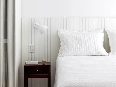 A minimalist bedroom featuring a white quilted blanket on a bed with a white headboard and wooden nightstand against a white