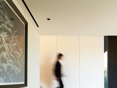 A modern home office with a wooden desk, two laptops, and a blurred person walking by the staircase.