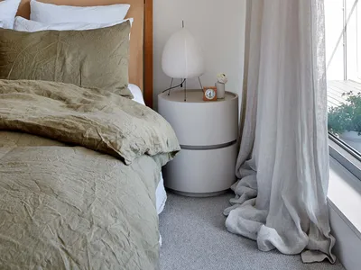 Modern minimalist bedroom featuring a bed with neutral bedding and a round bedside table with a lamp and alarm clock, alongsi