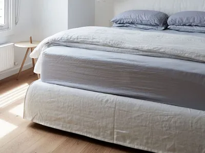 A minimalist bedroom with a bed dressed in light blue linen blankets and pillows, featuring a pendant light and a window with