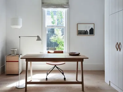 A minimalist home office featuring a wooden desk, modern chair, and metal cabinet seen through a window with greenery outside