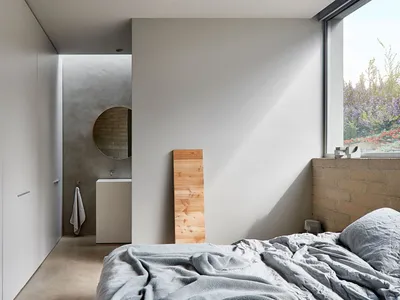 Minimalist bedroom with concrete walls and floor, featuring a bed and a small sink with a circular mirror.