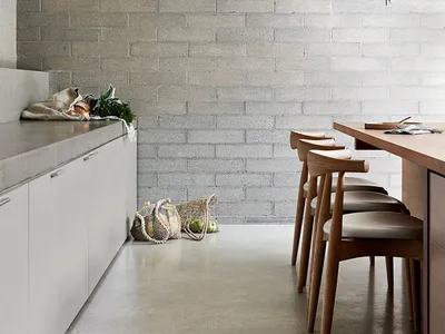 Modern minimalist kitchen with concrete countertops, white cabinets, and wooden stools against a textured brick wall.