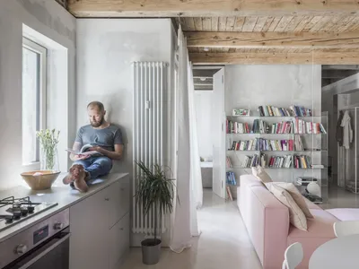 A minimalist living room with concrete and wood materials, featuring a pink sofa, bookshelf, and man reading.