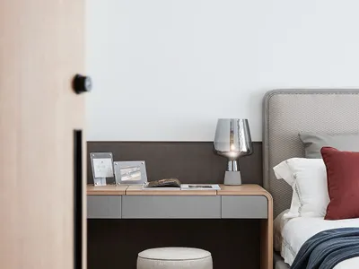 A minimalist bedroom featuring a grey wooden bed frame, soft grey carpet, and a modern wooden desk with a grey cushion.
