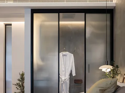 Bedroom featuring a white bed with a beige pillow, a glass sliding door with a white robe, and a plant in the corner.