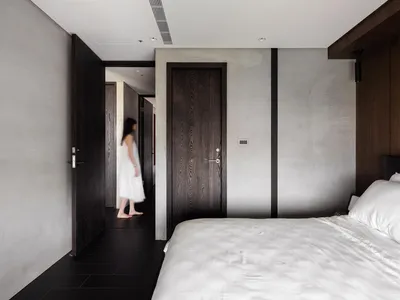A minimalist bedroom with concrete walls, a white bed, and a person walking through the hallway.