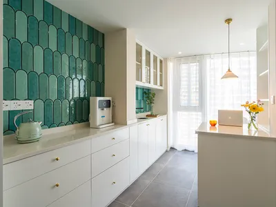 A modern, minimalist kitchen featuring green ceramic tiles, white cabinetry, and a white countertop with a kettle, laptop, an