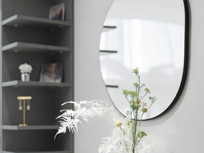 A modern home office with concrete and glass elements featuring a green sofa, a round mirror, a black vase with dried plants,