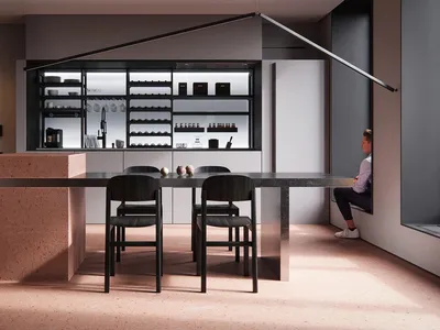 A modern, minimalist kitchen featuring a concrete island and table with black chairs, illuminated shelves, and a person sitti