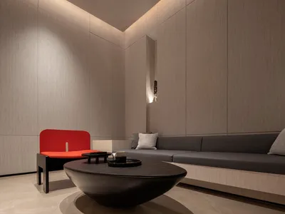 A minimalist living room with a curved concrete coffee table, a red accent chair, and a built-in gray sofa with patterned thr