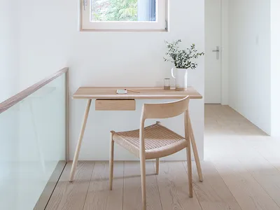 A minimalist home office with a wooden desk and chair, featuring a small window and natural lighting.