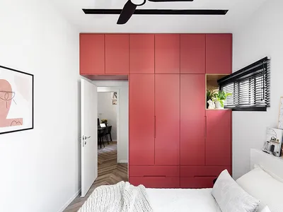 A modern minimalist bedroom featuring a large red cabinet, white bed, and white walls, with a view into a hallway with herrin
