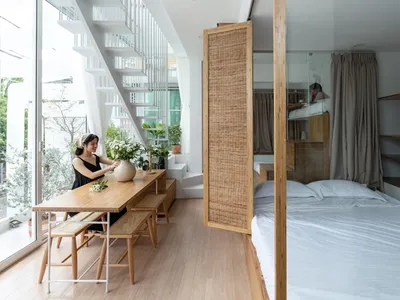 A modern minimalist bedroom featuring a large bed with white sheets, a wooden table with a vase and plants, and a large windo